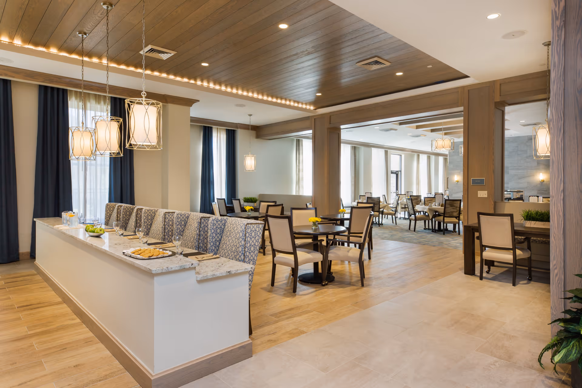 A spacious and well-lit dining area in a senior living facility featuring multiple round tables with chairs, a long counter with cushioned bench seating, pendant lights hanging from a wooden ceiling, and large windows with curtains allowing natural light to fill the room.