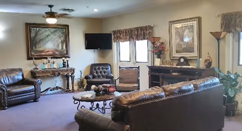 A cozy living room with brown leather sofas and armchairs arranged around a round coffee table. The room has beige walls adorned with framed artwork and a wall-mounted TV. There are two windows with valance curtains, a decorative console table with candles, and a floor lamp. A potted plant is visible in the corner.