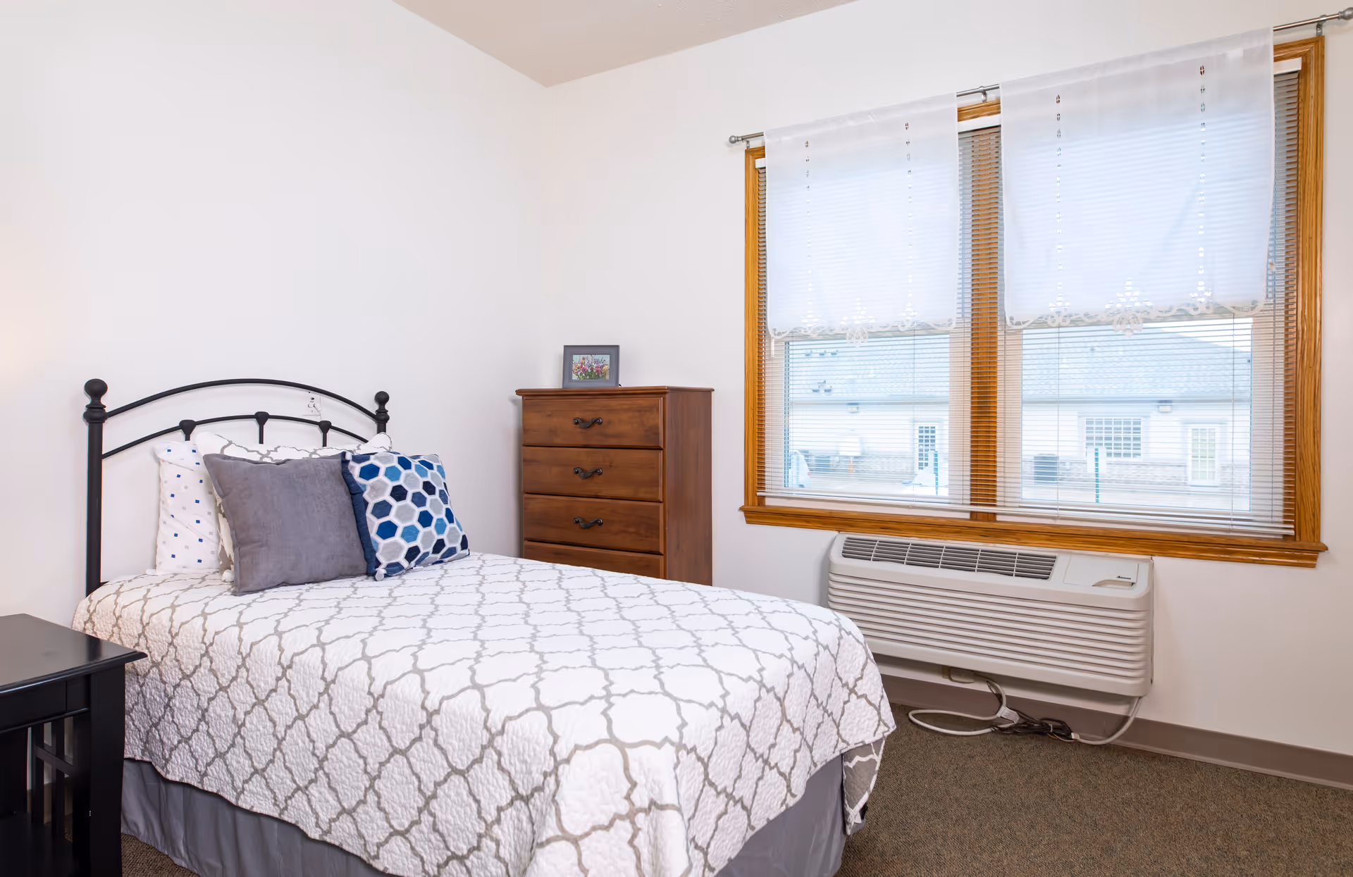 A neatly made single bed with a white and gray patterned bedspread and three pillows, positioned against a white wall. Next to the bed is a small black nightstand. Across from the bed is a wooden chest of drawers with a small framed picture on top. Two large windows with white blinds and sheer curtains let in natural light. Below the windows is a wall-mounted air conditioning unit.