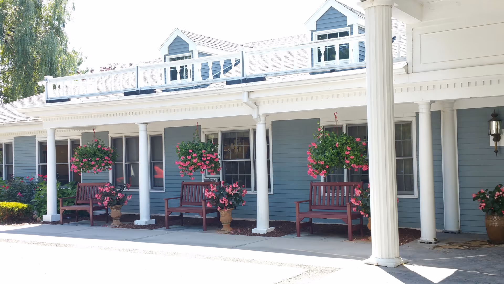 Front porch of a blue building with white columns, wooden benches, and hanging pink flower baskets.