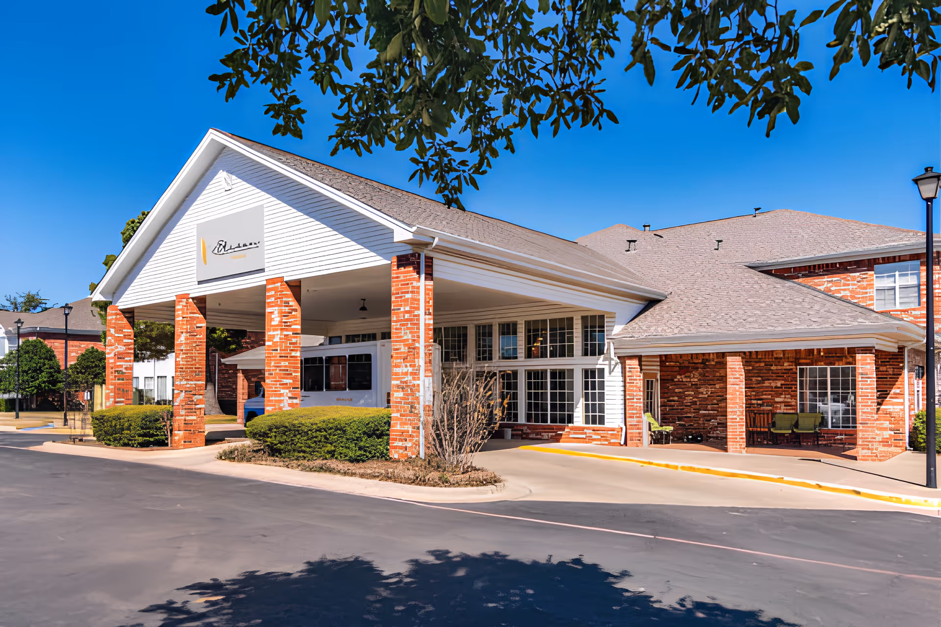 Covered front entrance and porte-cochère of a red-brick assisted living building with shrubs under a clear blue sky.