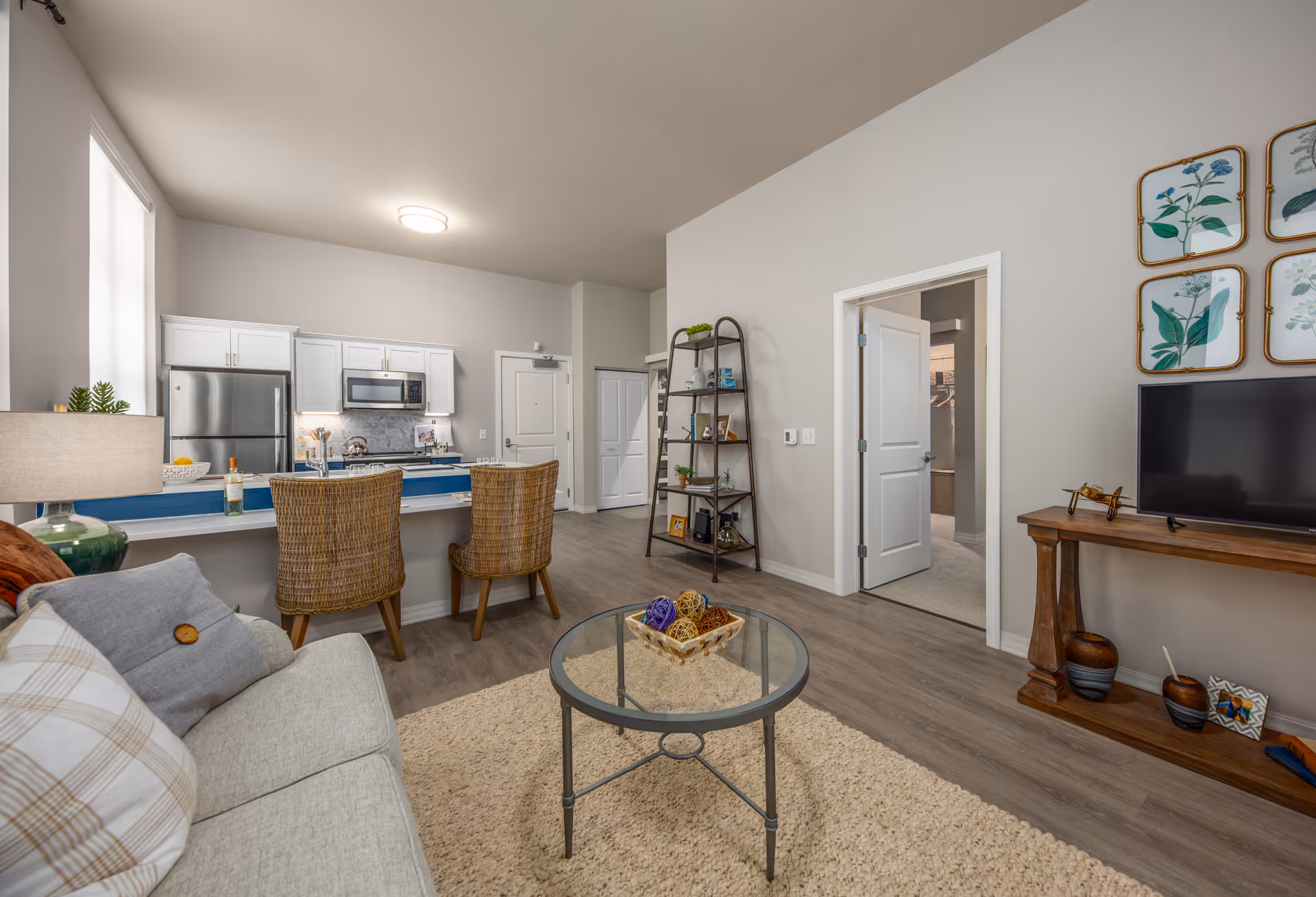 A bright and modern living area in a senior living facility featuring a light gray sofa with pillows, a glass coffee table with decorative items, and a wooden console table with a TV and decor. The open kitchen has white cabinets, stainless steel appliances, and a breakfast bar with two wicker chairs. The room has light wood flooring, neutral walls, and botanical artwork on the wall.