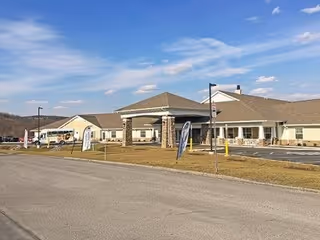 Exterior view of a single-story senior living facility building with a covered entrance supported by stone pillars, a driveway, and a parking area. The sky is partly cloudy and the surrounding area includes grass and hills in the background.
