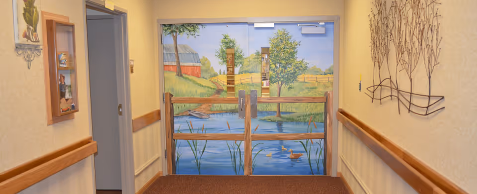A hallway in a senior living facility with beige walls and wooden handrails. At the end of the hallway, double doors are painted with a mural depicting a peaceful outdoor scene with a pond, ducks, trees, a red barn, and a wooden fence.