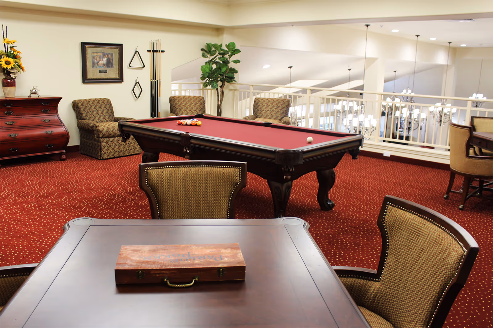 A cozy recreational room with a red carpet featuring a pool table with balls arranged for a game. There are several upholstered chairs around the room, a wooden chest of drawers with a vase of sunflowers on top, and a framed picture on the wall. The room overlooks a lower level with chandeliers visible through a white railing.