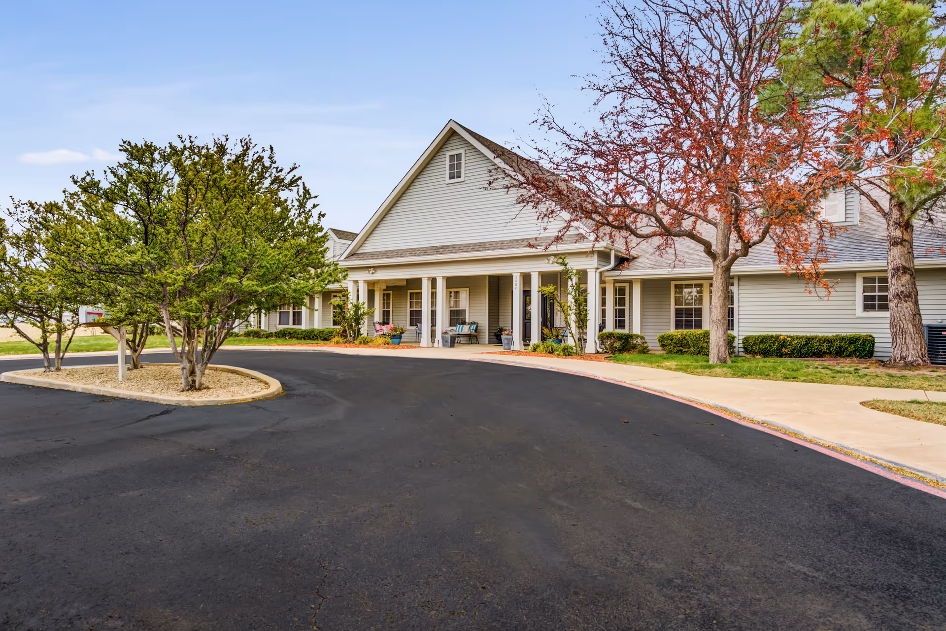 Front entrance of a single-story senior living building with a covered porch, driveway, and landscaped trees.