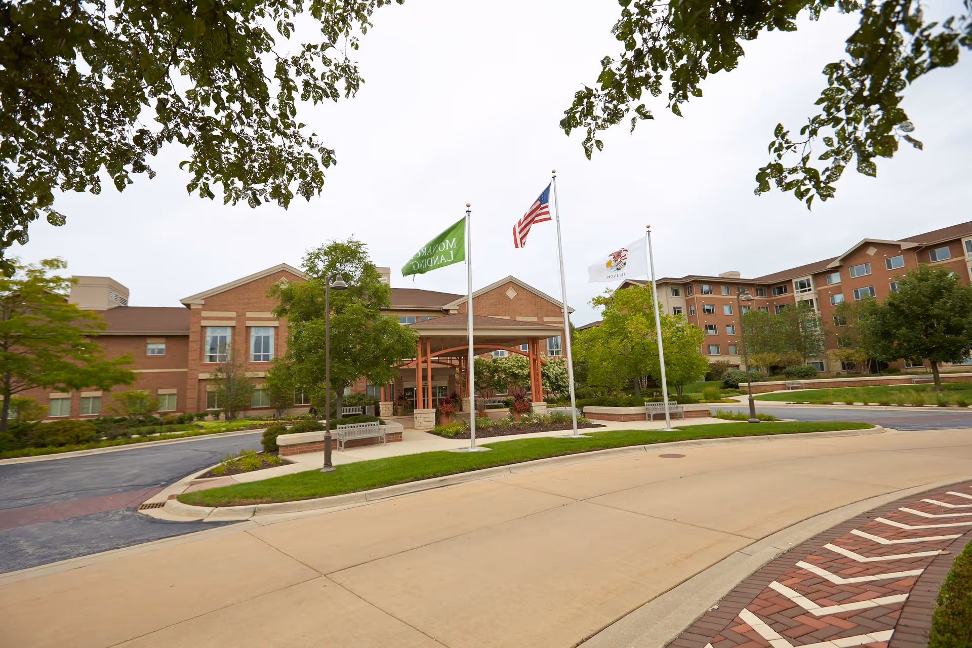 Exterior view of Monarch Landing senior living facility showing a large brick building with multiple windows, a covered entrance with three flagpoles flying the Monarch Landing flag, the American flag, and another flag. The foreground includes a curved driveway, landscaped greenery, benches, and trees framing the scene.