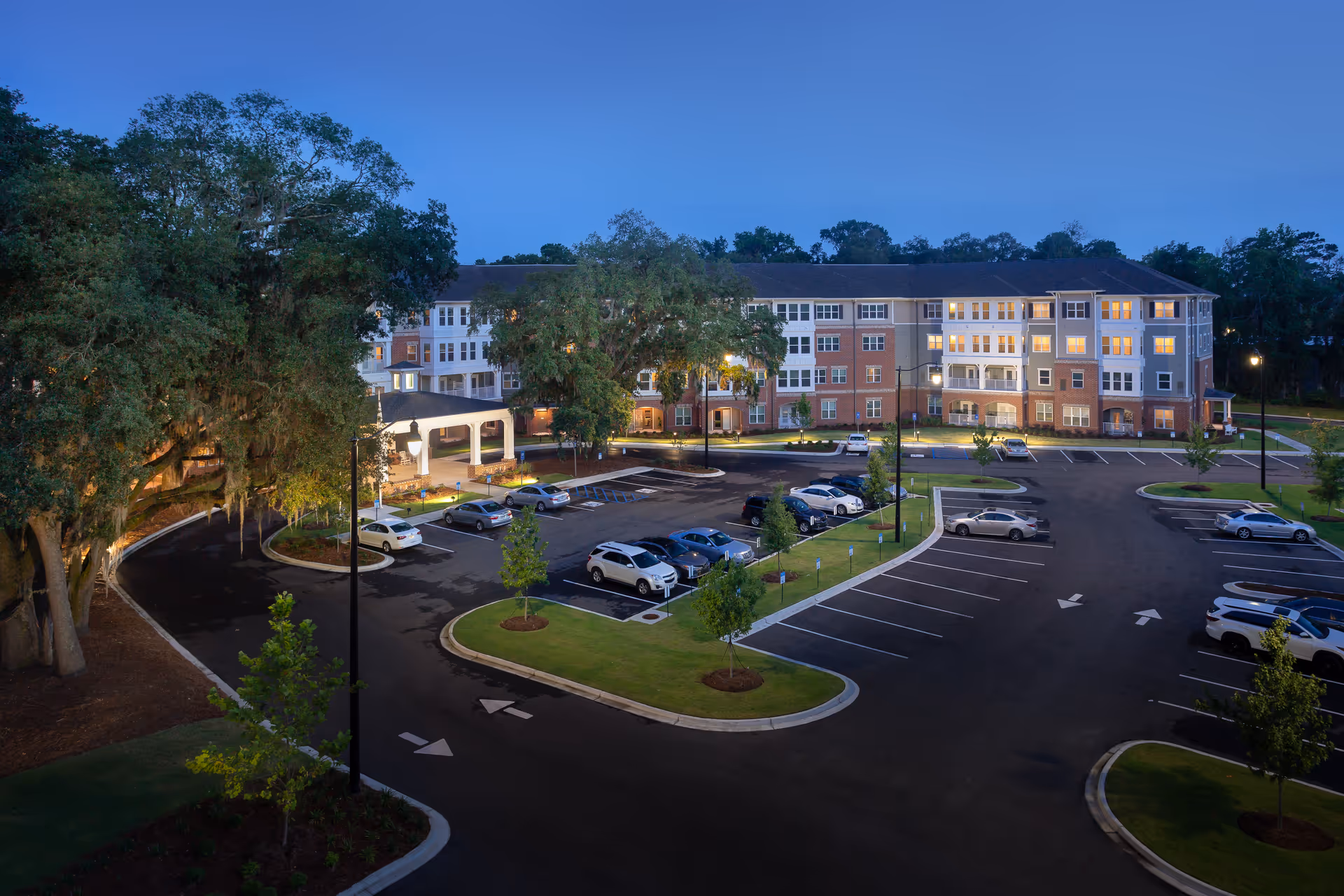 Exterior dusk view of the Harmony at Savannah senior living building with illuminated windows and a large parking lot in front.