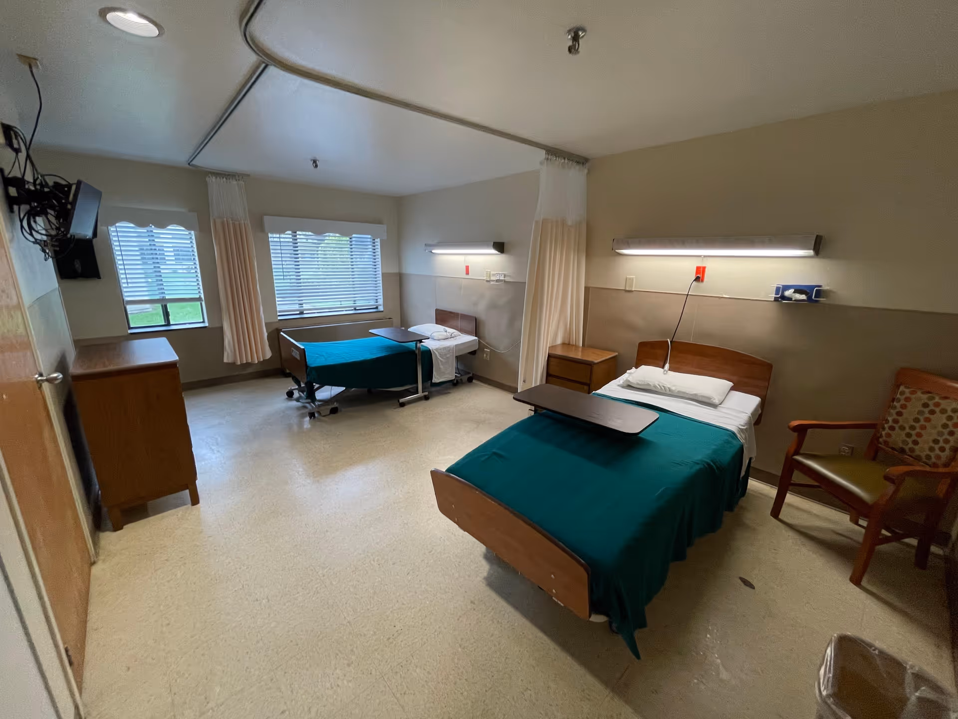 A room with two hospital-style beds each covered with green blankets and white pillows. Each bed has an overbed table. There is a wooden dresser, a wooden chair with a cushion, and windows with blinds and curtains. The room has beige walls and a tiled floor, with ceiling-mounted lights and a curtain track between the beds.