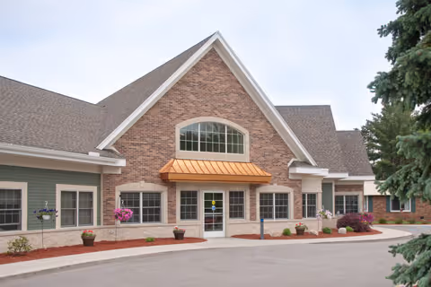 Front entrance of a brick and green-sided single-story building with a peaked roof, copper awning over glass doors, potted plants, and a driveway.