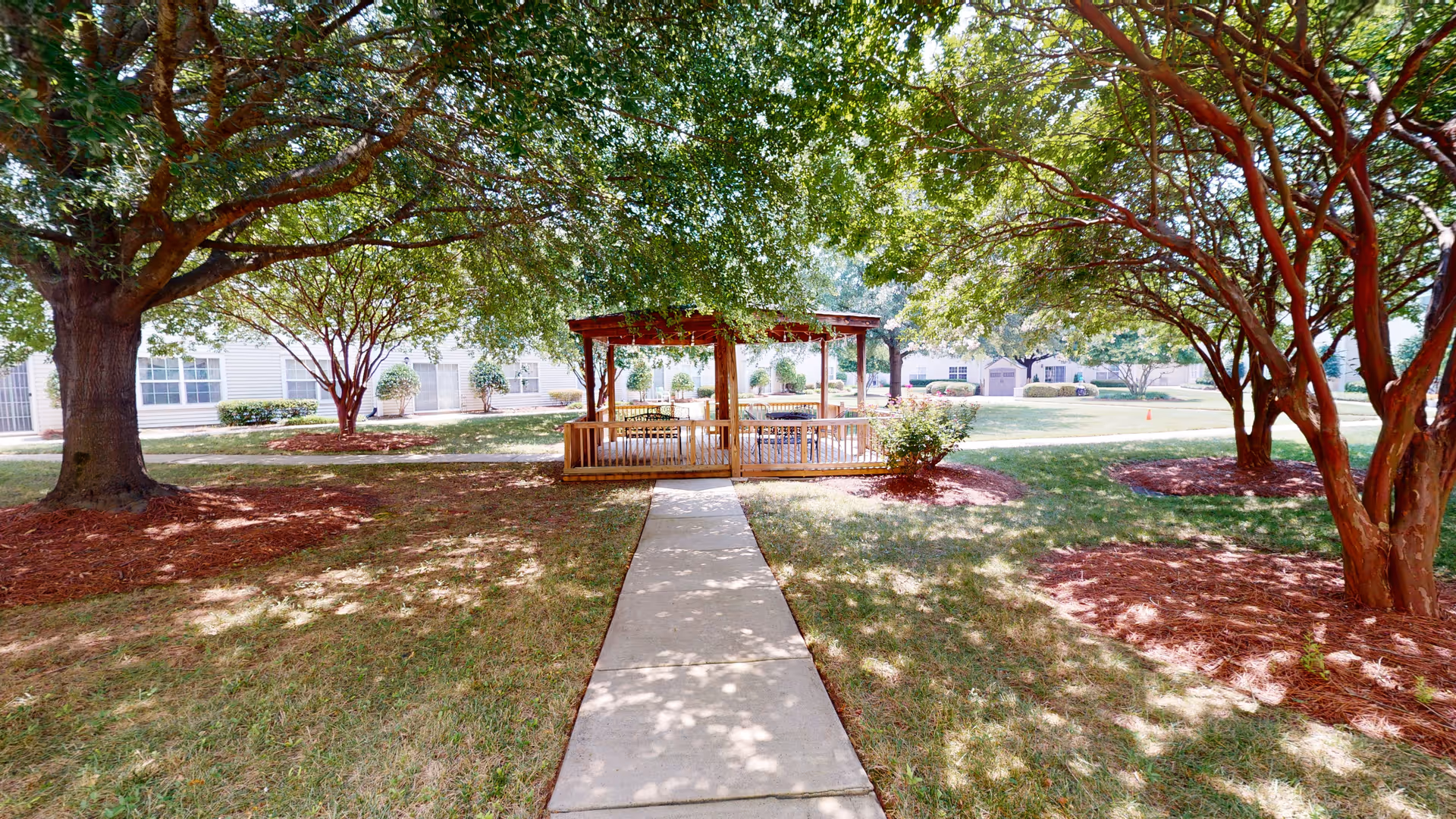 A shaded outdoor area with a wooden gazebo surrounded by trees and grass. A concrete pathway leads to the gazebo, which has benches inside. Residential buildings are visible in the background.