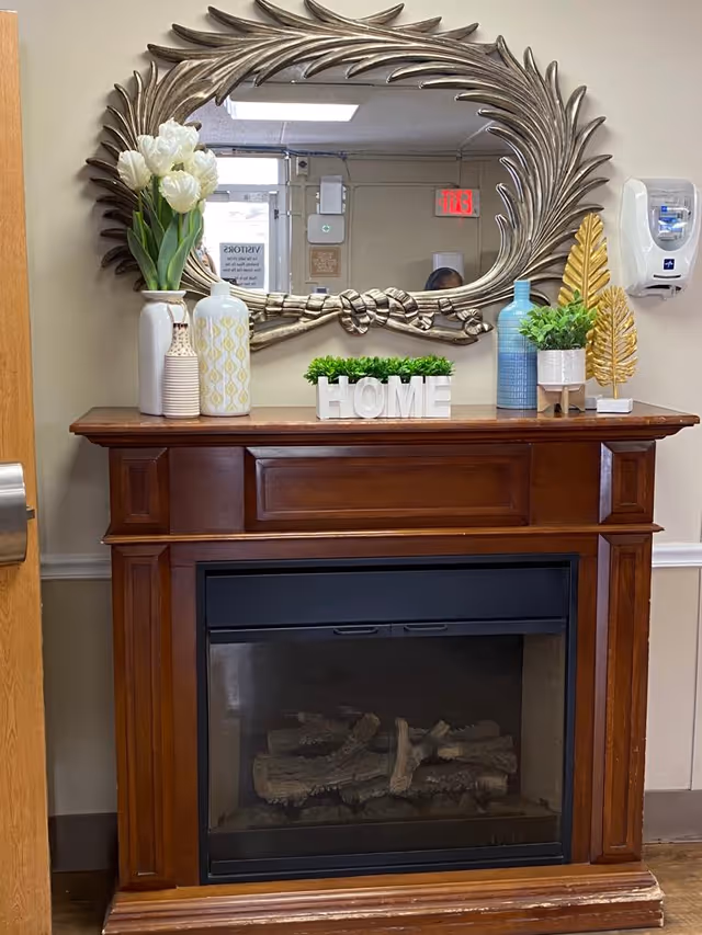 A wooden electric fireplace with decorative items on the mantel including white and blue vases, a small green plant, a decorative gold leaf, and white letters spelling HOME. Above the fireplace is an ornate mirror with a leaf design frame. A hand sanitizer dispenser is mounted on the wall to the right.