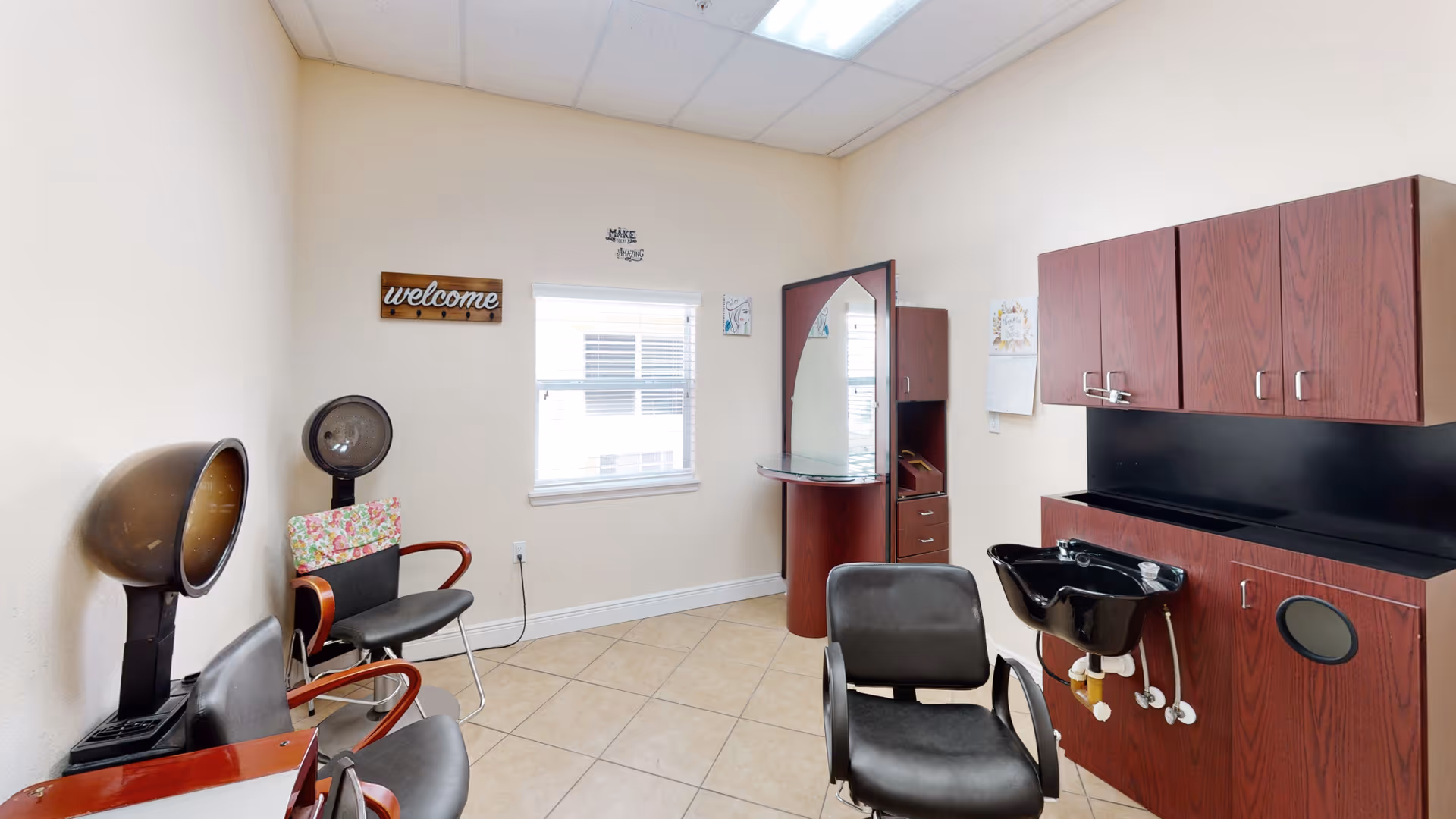 Small salon room with hooded hair dryers, styling chairs, a mirrored workstation with drawers, a shampoo sink, and wall cabinets.