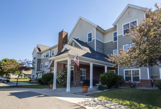 Exterior view of a multi-story senior living facility building with gray siding and brick accents. The entrance features a covered porch with white columns and an American flag hanging. There are trees, shrubs, and a paved driveway in front of the building under a clear blue sky.