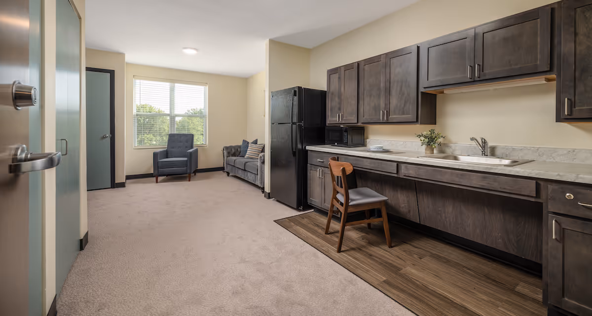 Spacious senior living studio with a kitchenette of dark cabinets and sink, a refrigerator, and a seating area with a sofa and chair by a window.