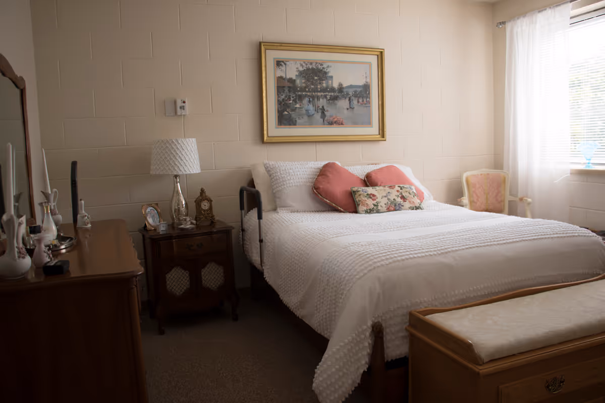 A cozy bedroom with a neatly made bed featuring white textured bedding and decorative pillows in pink and floral patterns. There is a wooden nightstand with a lamp, clock, and framed photo next to the bed. A wooden dresser with various decorative items and a mirror is on the left side. A cushioned bench is at the foot of the bed, and a window with sheer white curtains lets in natural light on the right side. A framed painting hangs on the wall above the bed.