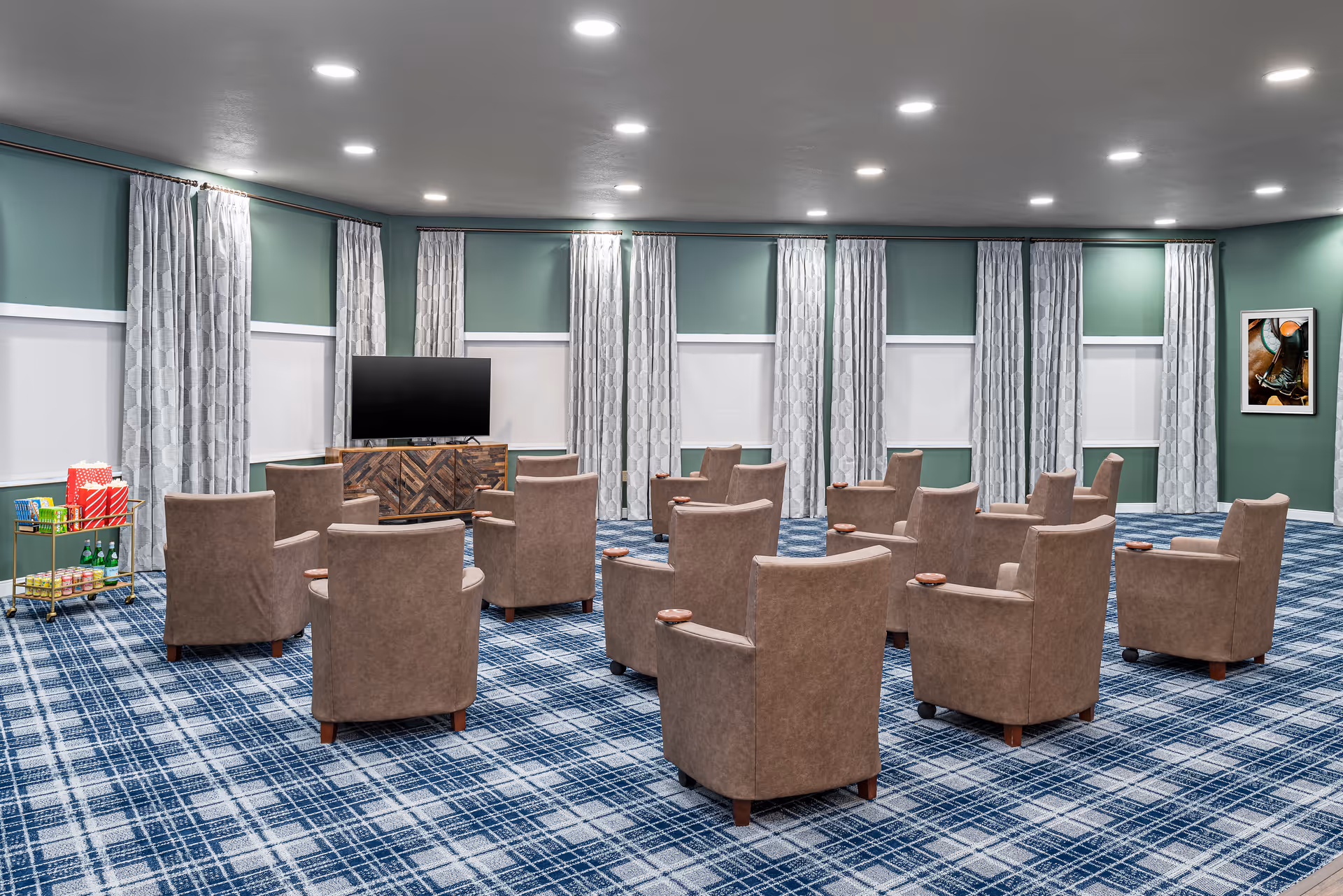 A spacious room with multiple brown armchairs arranged in rows facing a flat-screen TV on a wooden cabinet. The room has green walls with white trim, large windows covered with white blinds and patterned curtains, and a blue plaid carpet. A small cart with snacks and drinks is positioned on the left side of the room, and a framed picture hangs on the right wall.