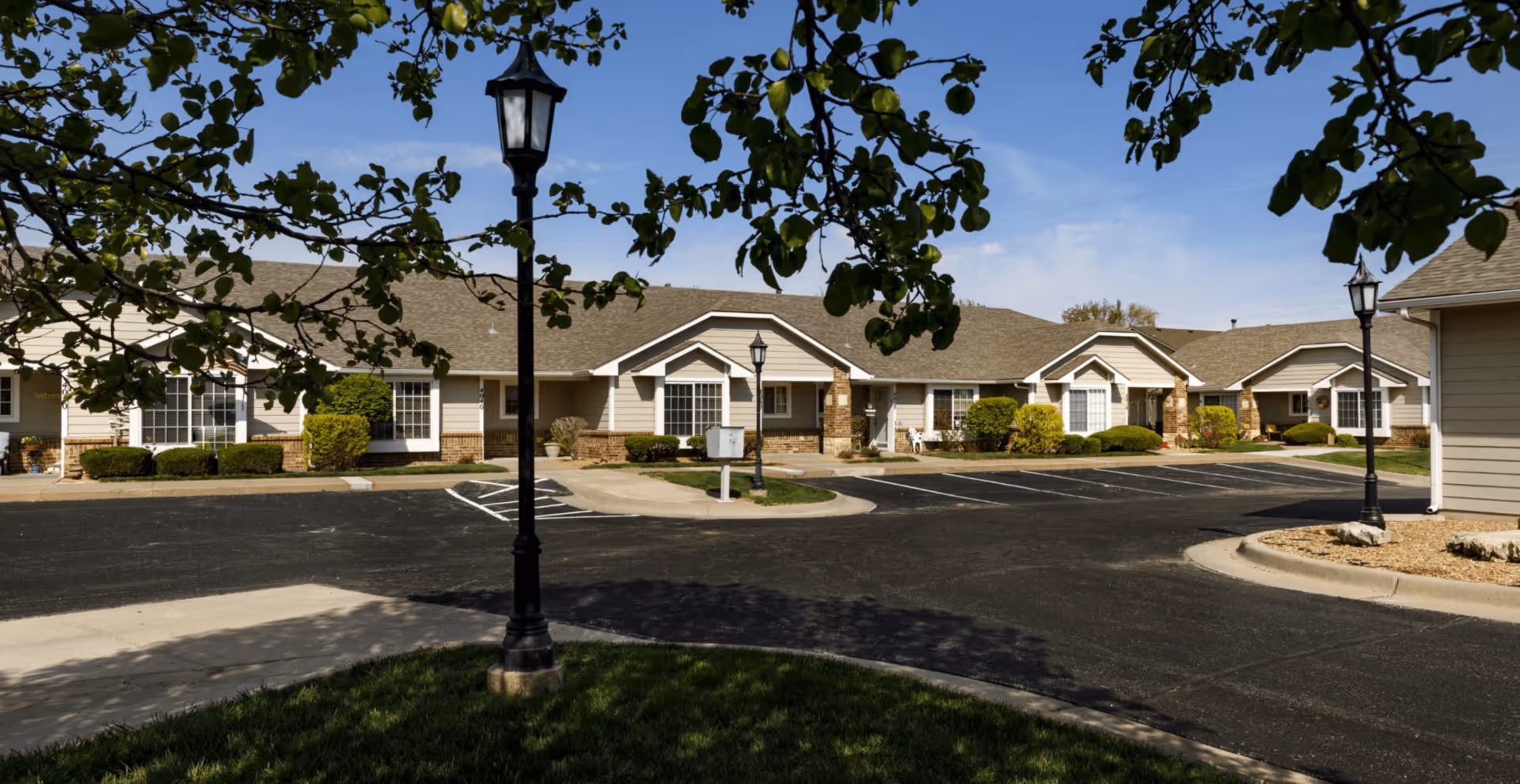 Exterior view of Homestead Estates of Wichita, showing a single-story building with beige siding and brick accents, surrounded by neatly trimmed bushes and trees, with a paved parking area and black lamp posts under a clear blue sky.