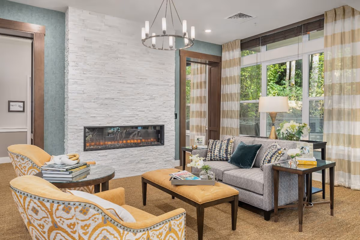 Contemporary living room with a gray sofa, patterned yellow armchairs, an ottoman, a linear fireplace set in a white stone wall, and large windows with striped curtains.