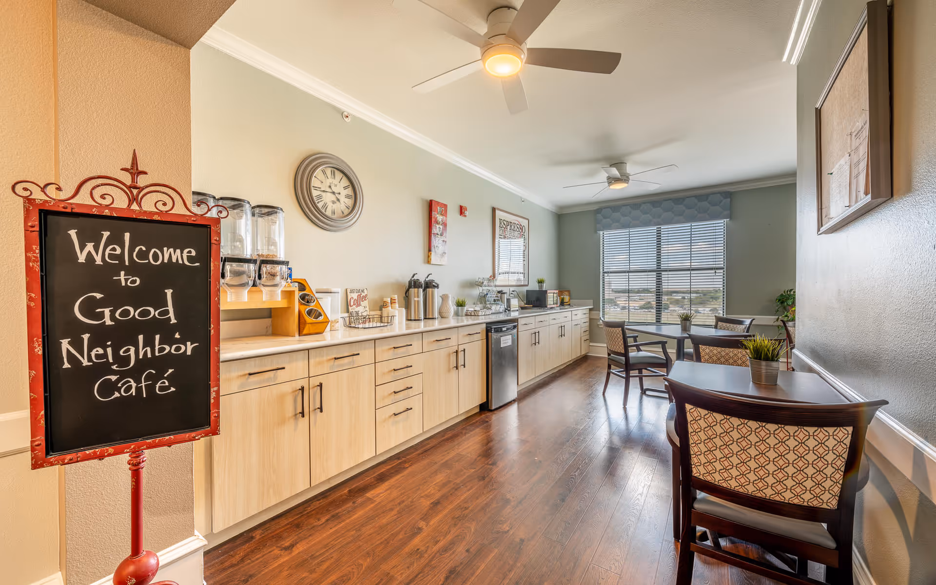 Interior view of a café area in a senior living facility with a sign that reads 'Welcome to Good Neighbor Café'. The room features a long counter with coffee dispensers, a microwave, and various coffee supplies. There are several tables and chairs arranged near a large window with blinds, and ceiling fans provide lighting and air circulation.