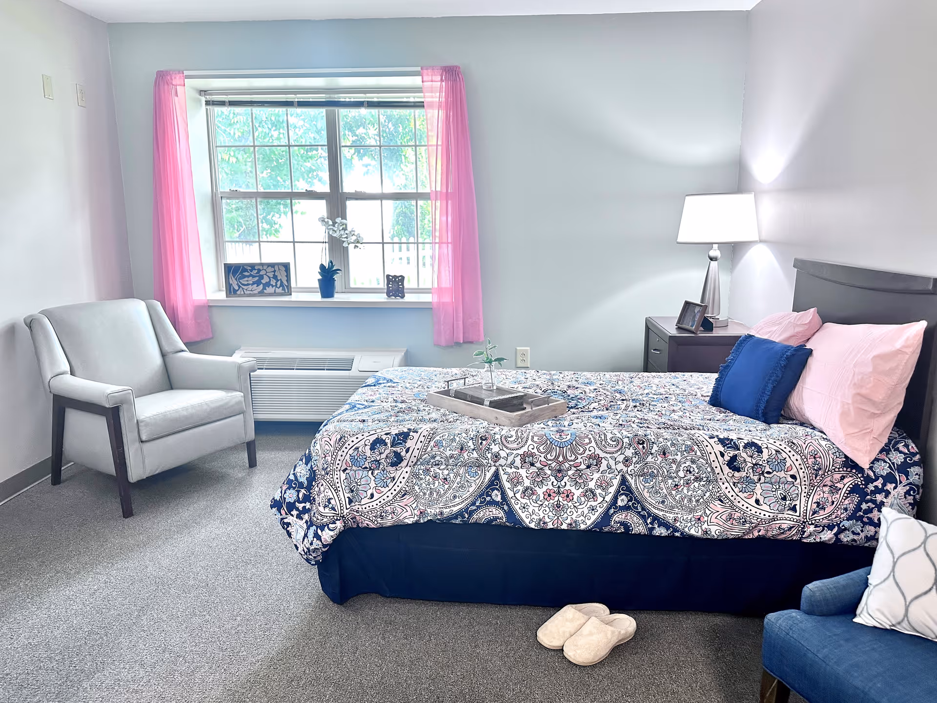 Well-lit bedroom with a bed dressed in patterned bedding, a nightstand and lamp, two armchairs, and a window with pink curtains.
