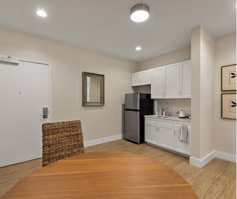 Small kitchenette with white cabinets, a stainless refrigerator and sink, and a round wooden dining table in the foreground.