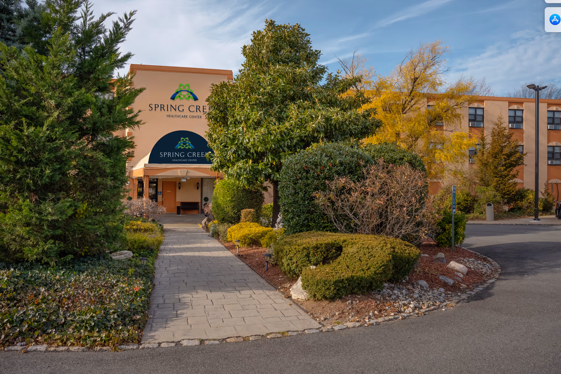 Exterior view of Spring Creek Healthcare Center showing the entrance pathway lined with green bushes and trees under a partly cloudy sky.
