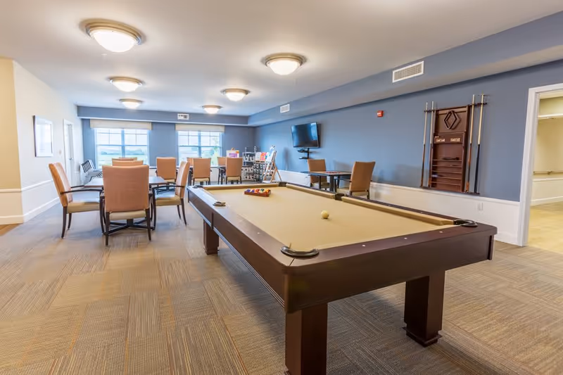 A spacious recreational room with a pool table in the foreground, several tables and chairs arranged near large windows, a wall-mounted TV, and a rack holding pool cues on a blue accent wall.