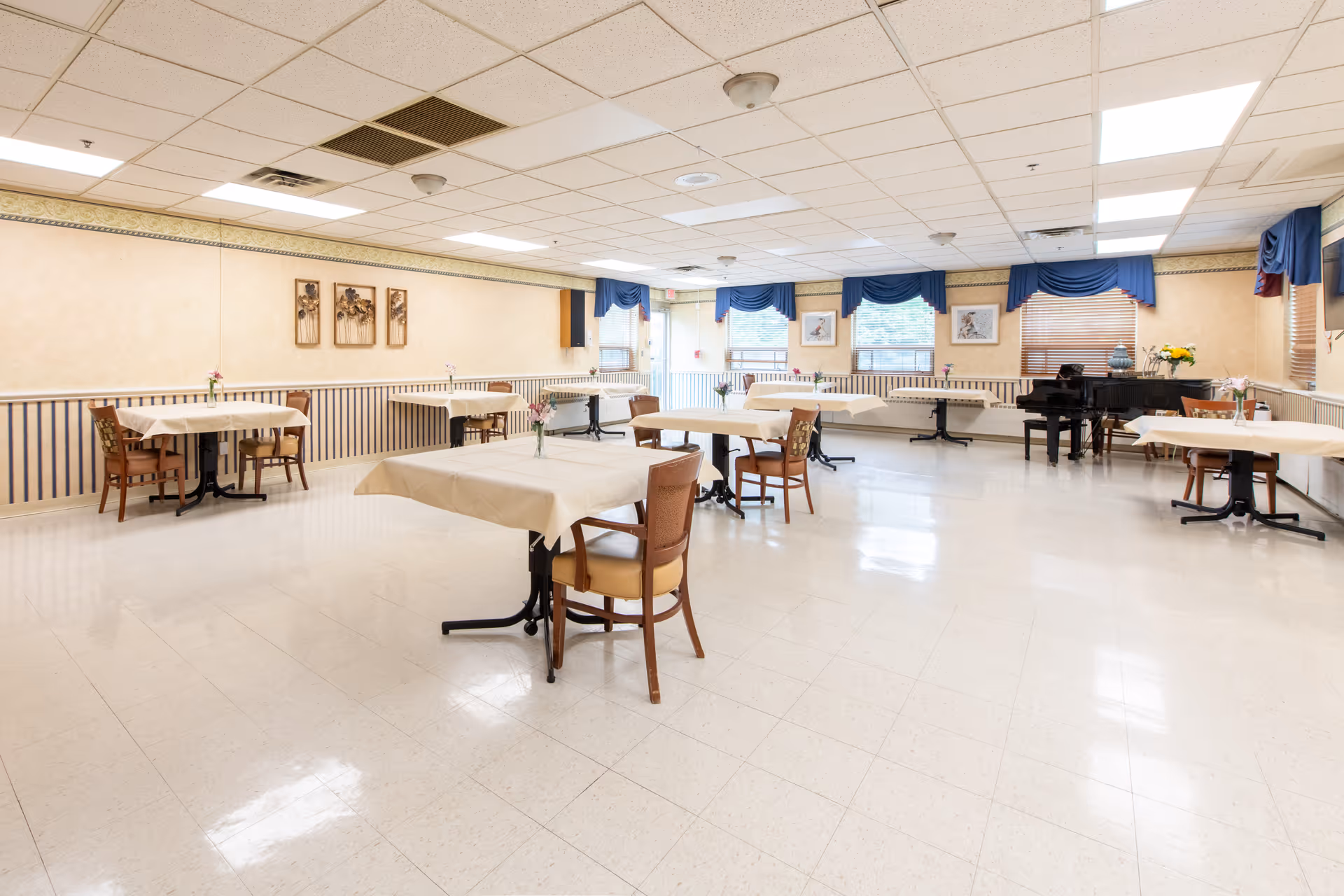 A spacious dining room with several tables covered with beige tablecloths, each accompanied by wooden chairs. The room has large windows with blue valances, framed artwork on the walls, and a black grand piano in the corner. The floor is tiled, and the ceiling has recessed lighting and ceiling fixtures.
