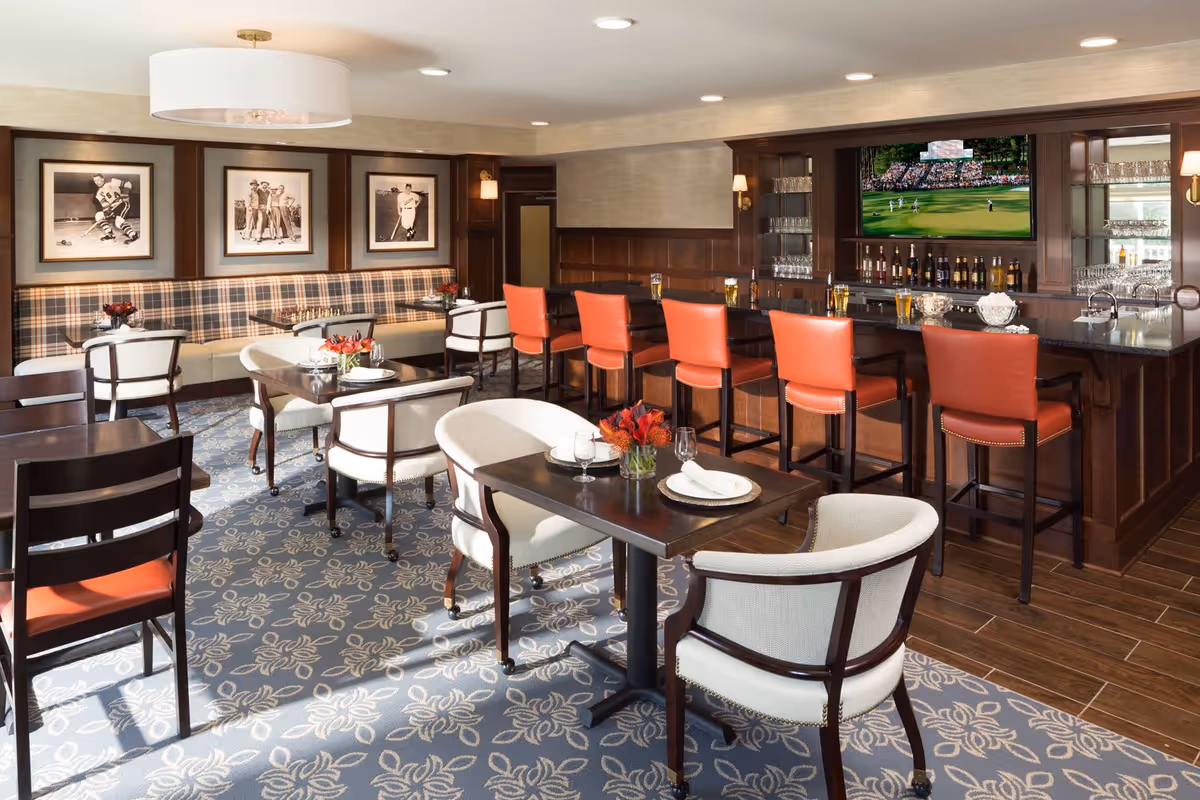 A cozy dining and bar area with several tables and chairs, a long bar counter with orange bar stools, and a large TV mounted above the bar showing a golf game. The room features patterned carpet, wood paneling, framed black and white sports photos on the wall, and soft lighting from ceiling fixtures and wall sconces.
