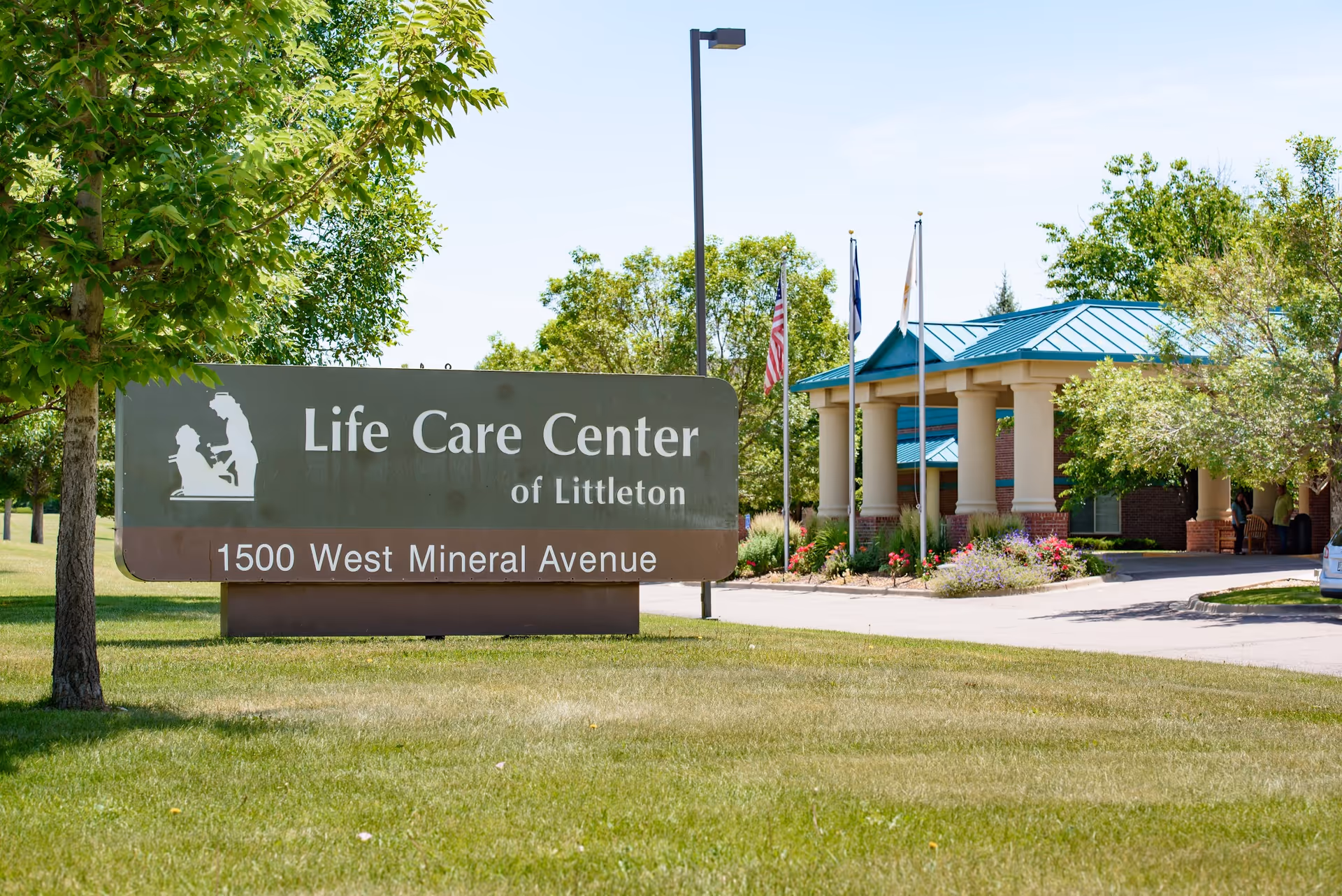 Entrance sign reading "Life Care Center of Littleton, 1500 West Mineral Avenue" with the facility entrance, flagpoles, and landscaping in the background.