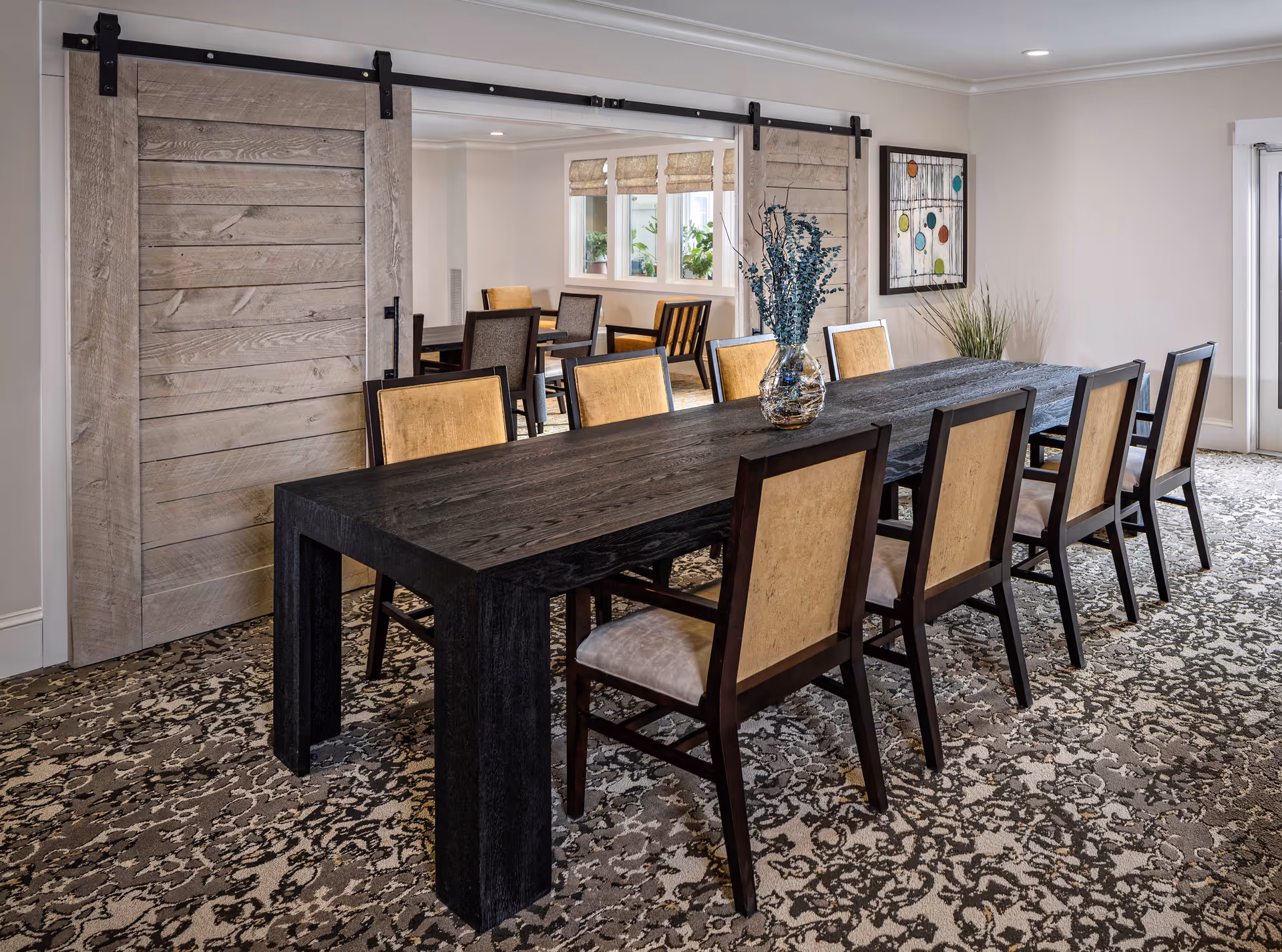 A spacious dining room with a long dark wooden table surrounded by eight chairs with light-colored upholstery. The room features patterned carpet flooring, two large sliding barn doors made of light wood, a decorative vase with blue foliage on the table, a framed abstract artwork on the wall, and windows letting in natural light.
