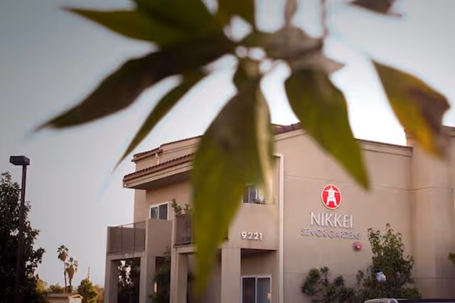 Exterior front of the Nikkei Senior Gardens building with the sign visible and blurred leaves in the foreground.