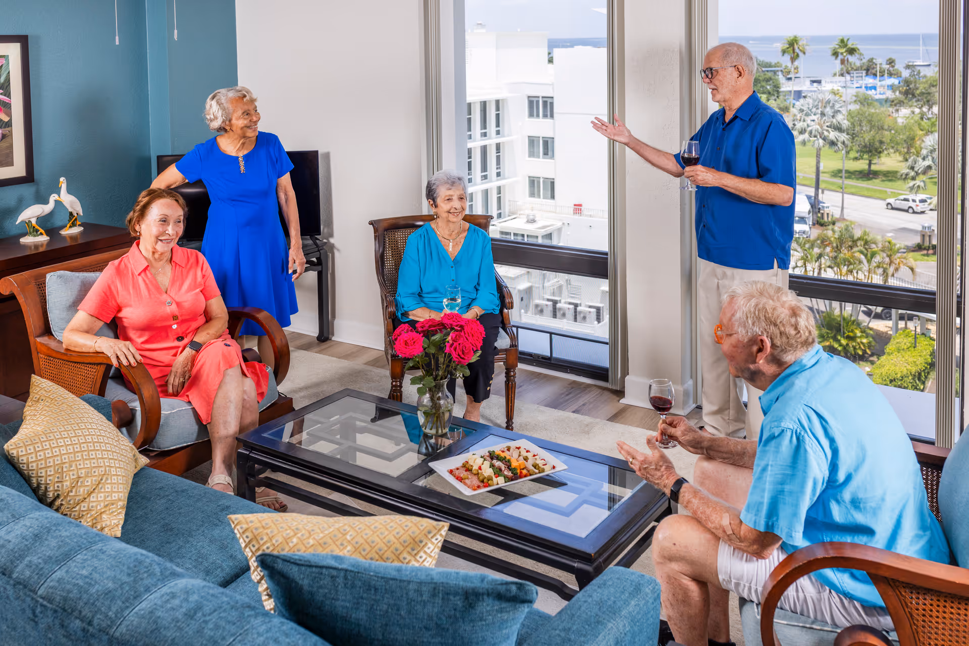 Five elderly people socializing in a bright living room with large windows showing a view of palm trees and a marina. Three women and two men are seated or standing around a glass coffee table with a vase of red roses and a plate of appetizers. Two men are holding glasses of red wine.