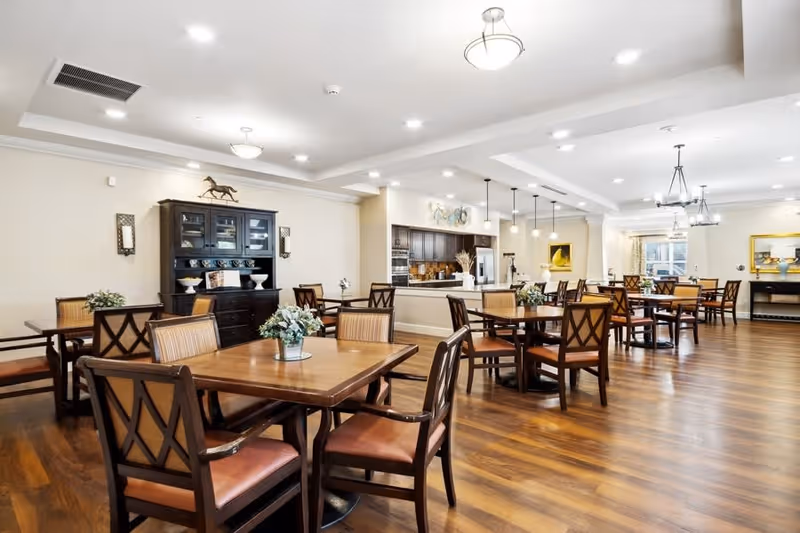 A spacious dining room with multiple wooden tables and chairs arranged neatly on a polished wooden floor. The room features warm lighting from ceiling fixtures, a dark wooden cabinet against the wall, and a kitchen area with dark cabinetry and pendant lights in the background. Small floral centerpieces decorate the tables.