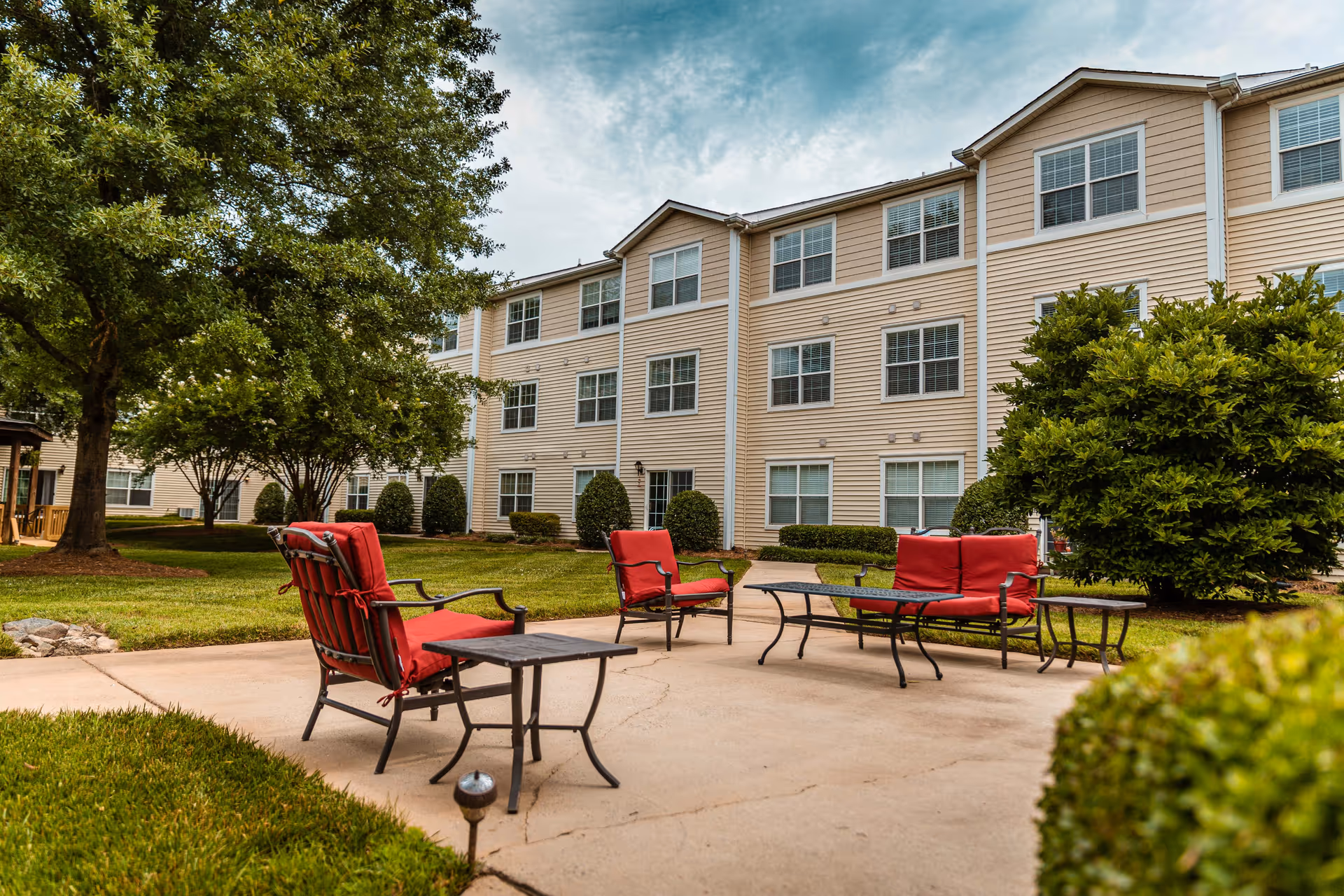 Outdoor patio with red-cushioned chairs and tables in front of a three-story beige residential building and landscaped lawn.