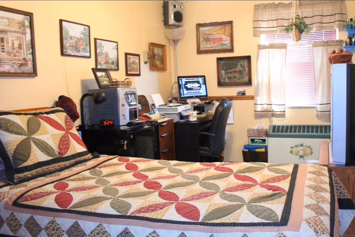 Cozy bedroom with a patterned quilted bed, desk with a computer and chair, framed wall art, and a curtained window.