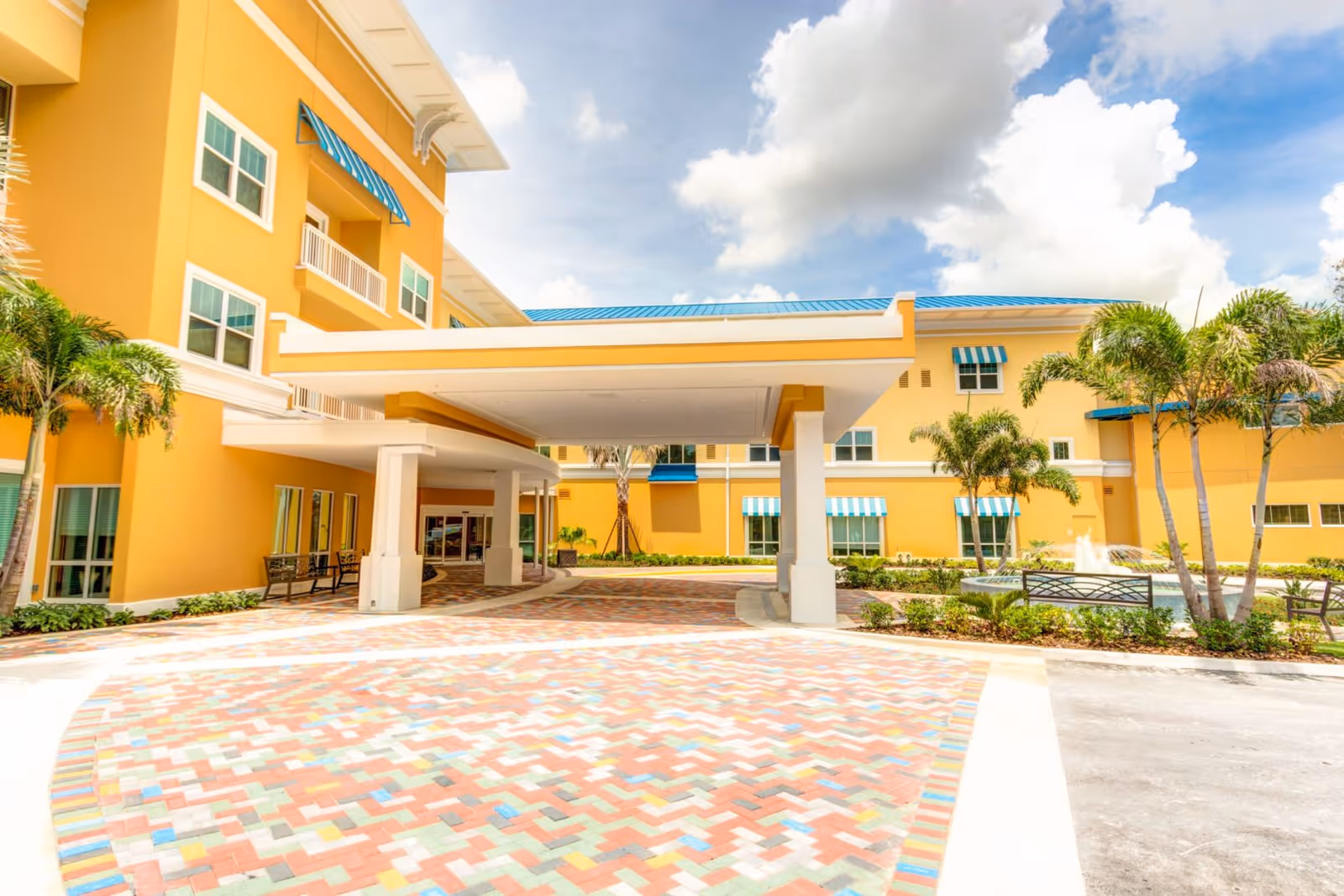 Entrance area of a senior living facility with a covered drop-off zone, colorful brick pavement, yellow building walls, blue and white striped window awnings, palm trees, benches, and a small fountain under a partly cloudy sky.