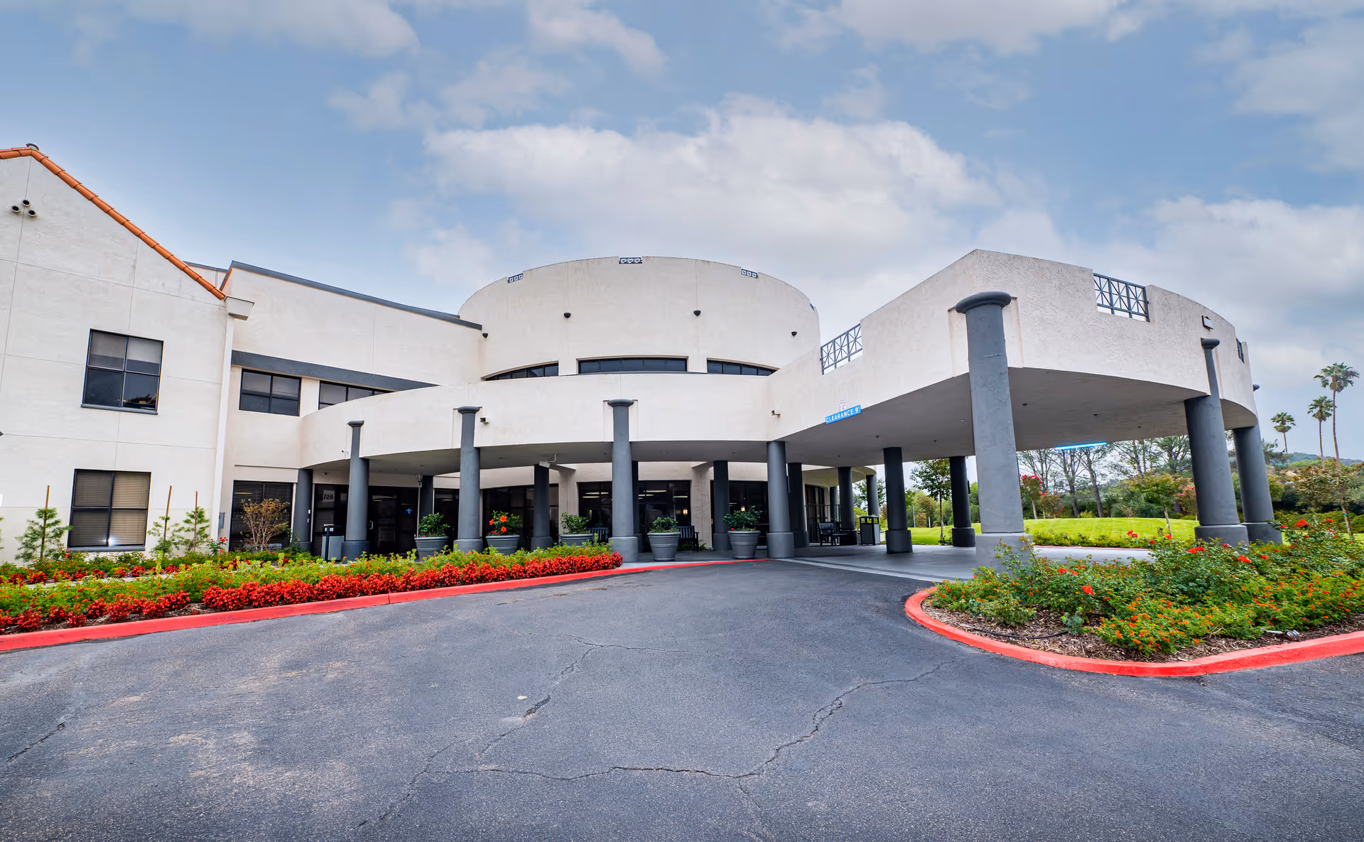 Exterior view of Villa Rancho Bernardo Care Center showing a two-story building with a covered entrance supported by multiple columns. The driveway is bordered by red curbs and landscaped with green bushes and red flowers. The sky is partly cloudy.