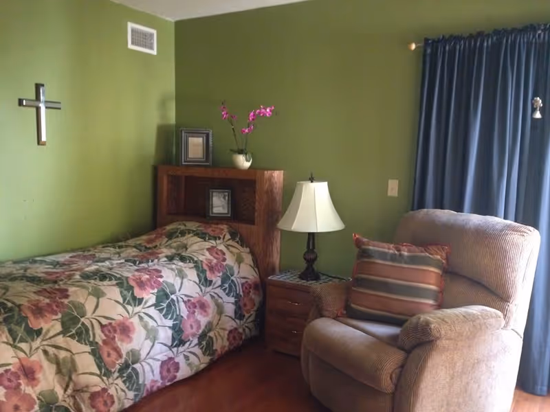 A cozy bedroom with green walls featuring a single bed with floral bedding, a wooden headboard with framed pictures and a vase with pink flowers, a bedside table with a lamp, and a comfortable armchair with a striped cushion next to a window with blue curtains.