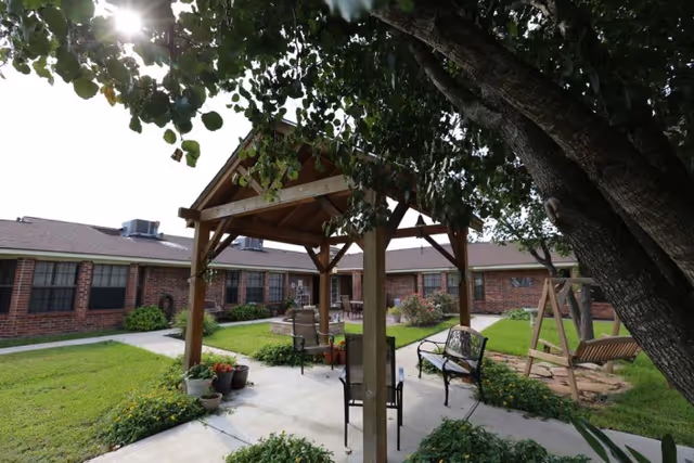 Outdoor courtyard area at Timberwood Nursing & Rehabilitation Center featuring a wooden gazebo with chairs underneath, surrounded by green grass, plants, and a large tree providing shade. There is also a wooden swing and brick building in the background.