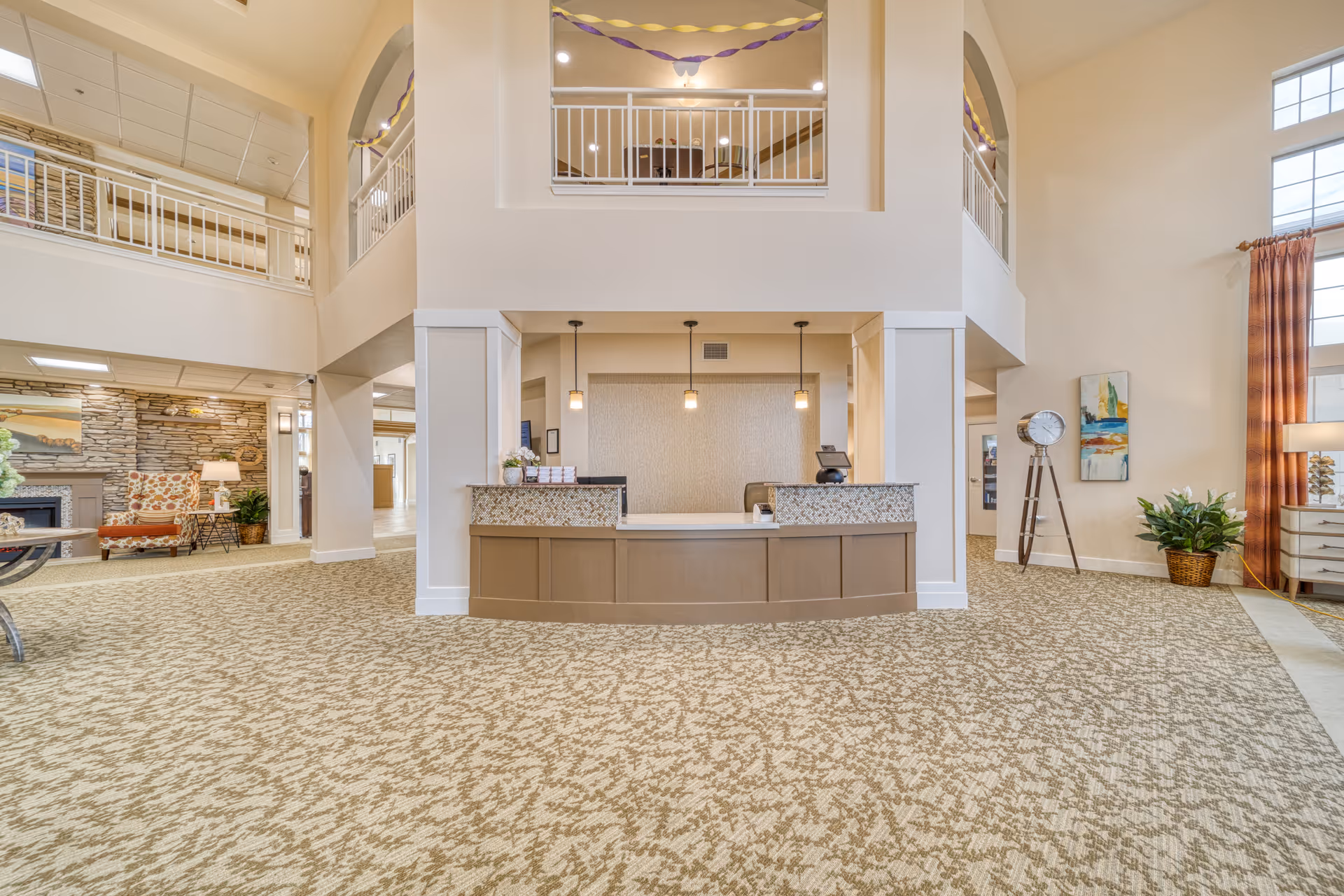Spacious senior living facility lobby with a reception desk in the center, beige patterned carpet, high ceilings, and a second-floor balcony. The area is decorated with plants, a standing clock, artwork, and comfortable seating near a stone fireplace.