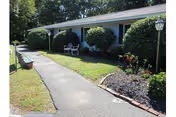 Single-story residential building with a paved walkway, manicured shrubs, and outdoor chairs near the entrance.