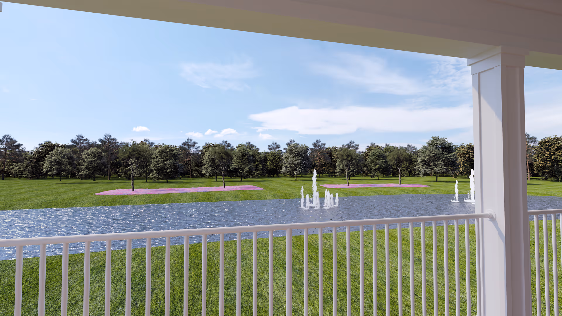 View from a covered porch with white railing overlooking a pond with multiple water fountains, surrounded by green grass, trees, and patches of purple flowers under a partly cloudy sky.