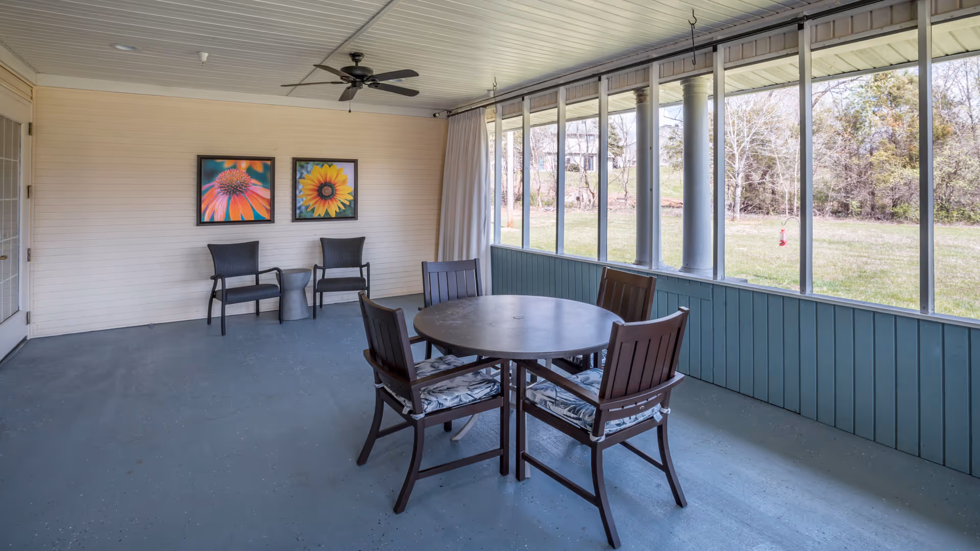A screened sunroom with a round table and four chairs, two additional chairs under two floral paintings, and large windows overlooking a grassy yard.