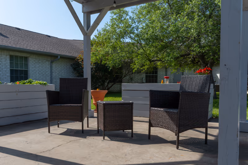 Three wicker chairs and a small wicker table on a shaded patio with planters and a building in the background.