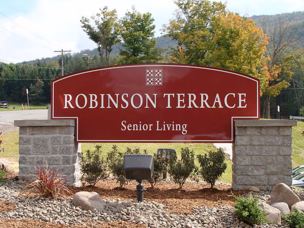 Red roadside sign reading "Robinson Terrace Senior Living" mounted between stone pillars with landscaping and trees behind.