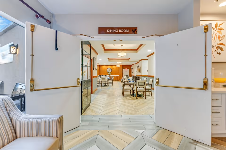View through double doors into a dining room with several tables and chairs arranged neatly. The floor has a light wood pattern, and the walls are decorated with wood paneling and framed artwork. A clock is visible on the far wall, and ceiling lights provide warm illumination.