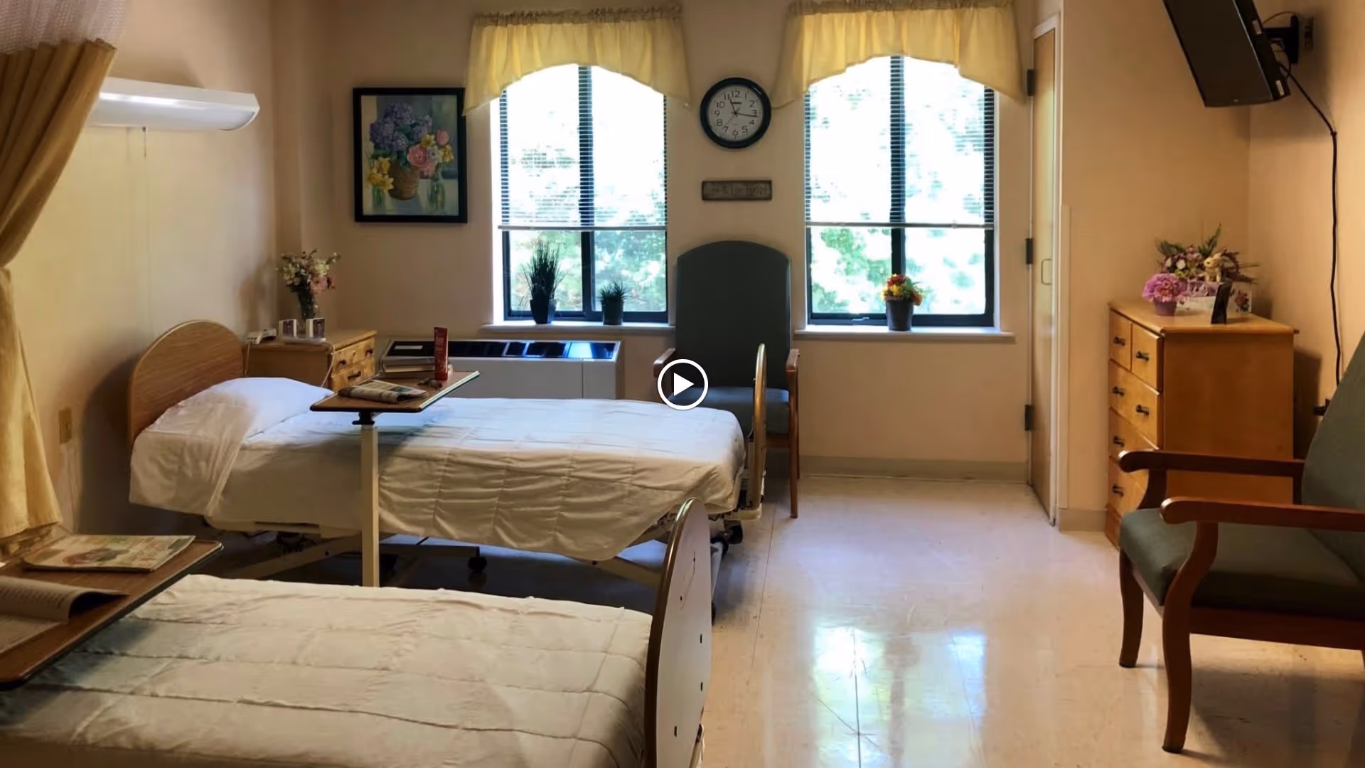 A bright and clean room with two hospital-style beds, each with white bedding and adjustable overbed tables. The room has two large windows with yellow valances, a clock on the wall between the windows, and several potted plants on the windowsills. There are two wooden dressers, a green upholstered chair, and a wall-mounted television on the right side. The floor is light-colored and shiny.