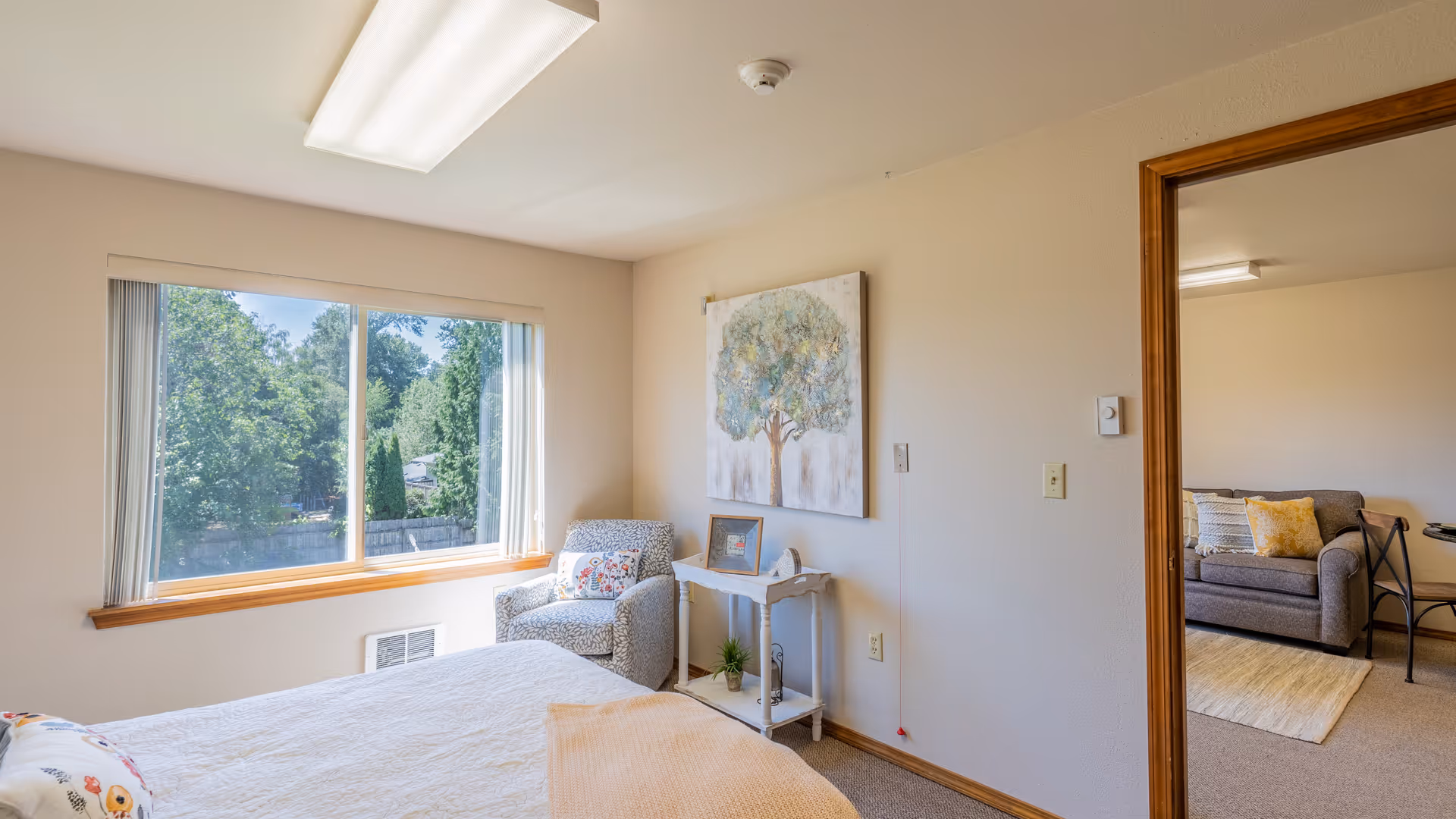 A bright bedroom with a large window showing green trees outside. The room has a bed with a white quilt and a peach blanket, a patterned armchair with a decorative pillow, a small white side table with a plant and picture frame, and a painting of a tree on the wall. An open doorway leads to a living room area with a gray sofa and yellow and white pillows.